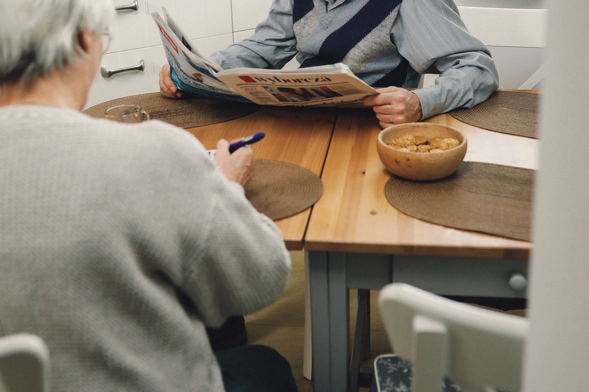 Un couple de retraités à table : lui lit le journal, elle fait des mots croisés.